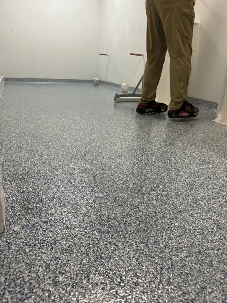 Worker installing commercial flake epoxy flooring in a large room with gray and white speckled pattern
