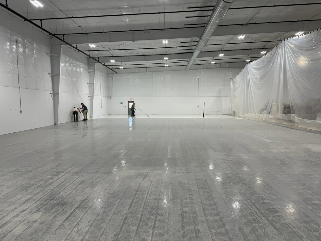Workers applying a concrete floor coating in a large commercial warehouse with plastic sheeting containment