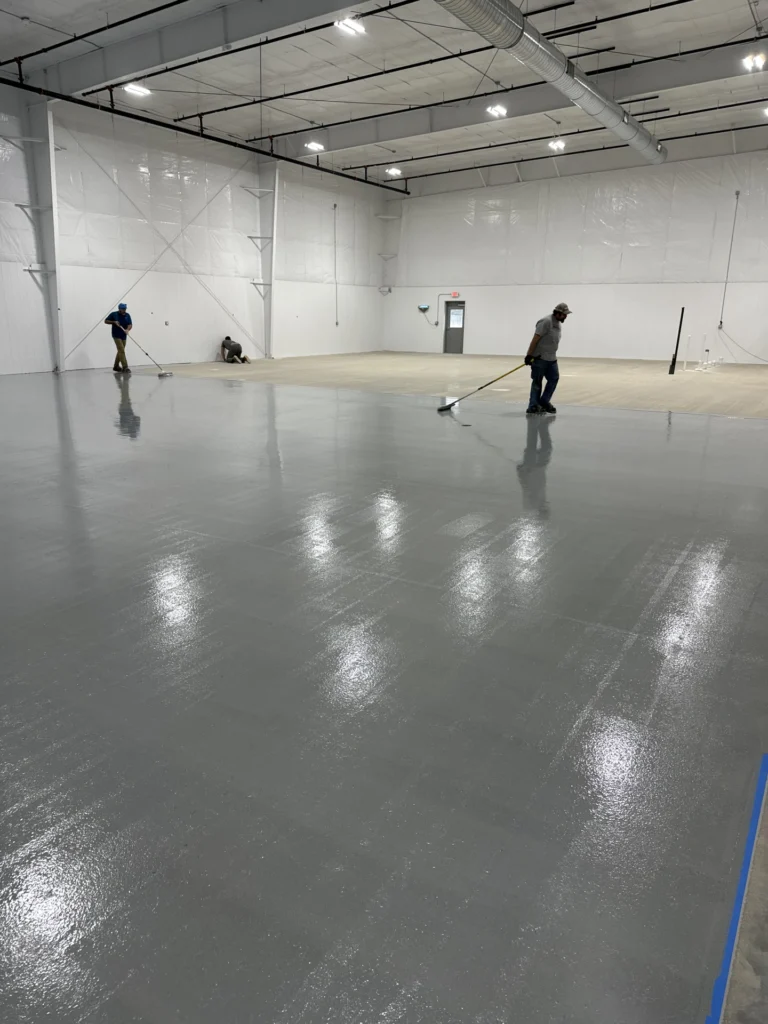 Workers applying a smooth, glossy floor coating in a large commercial warehouse with high ceilings and bright lighting