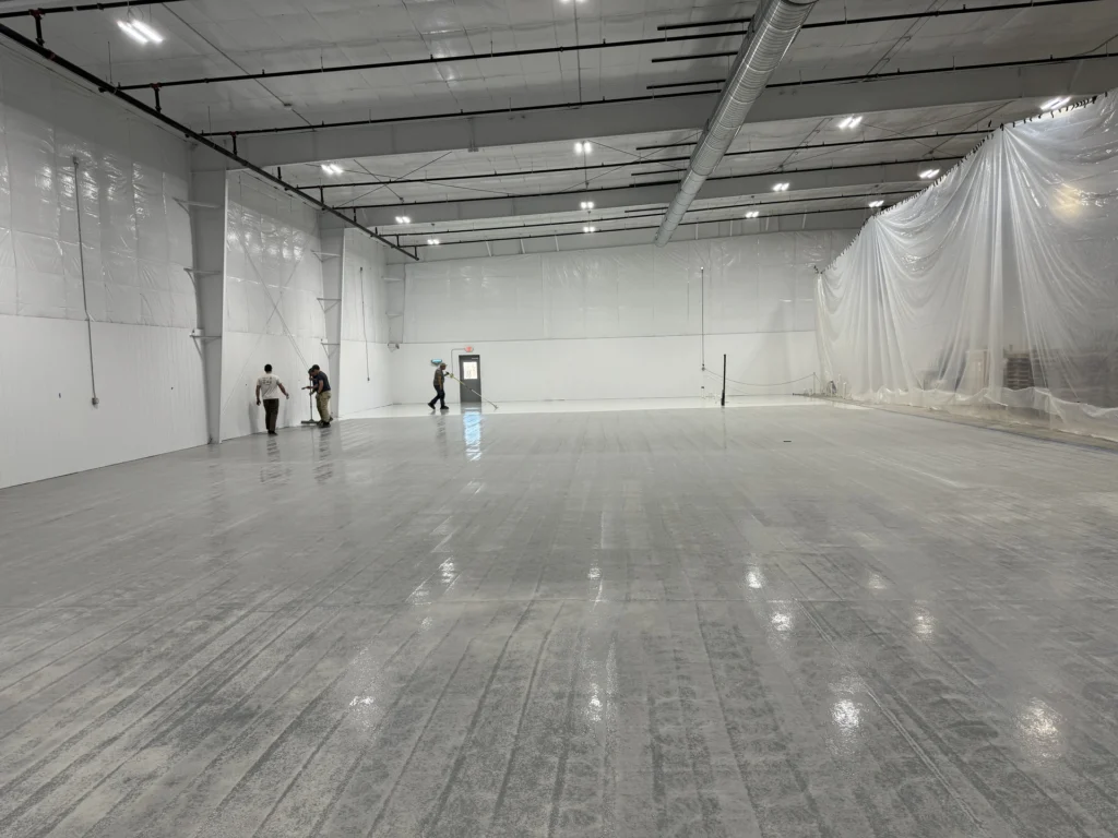 Workers installing a smooth, glossy floor coating in a large commercial warehouse with plastic sheeting