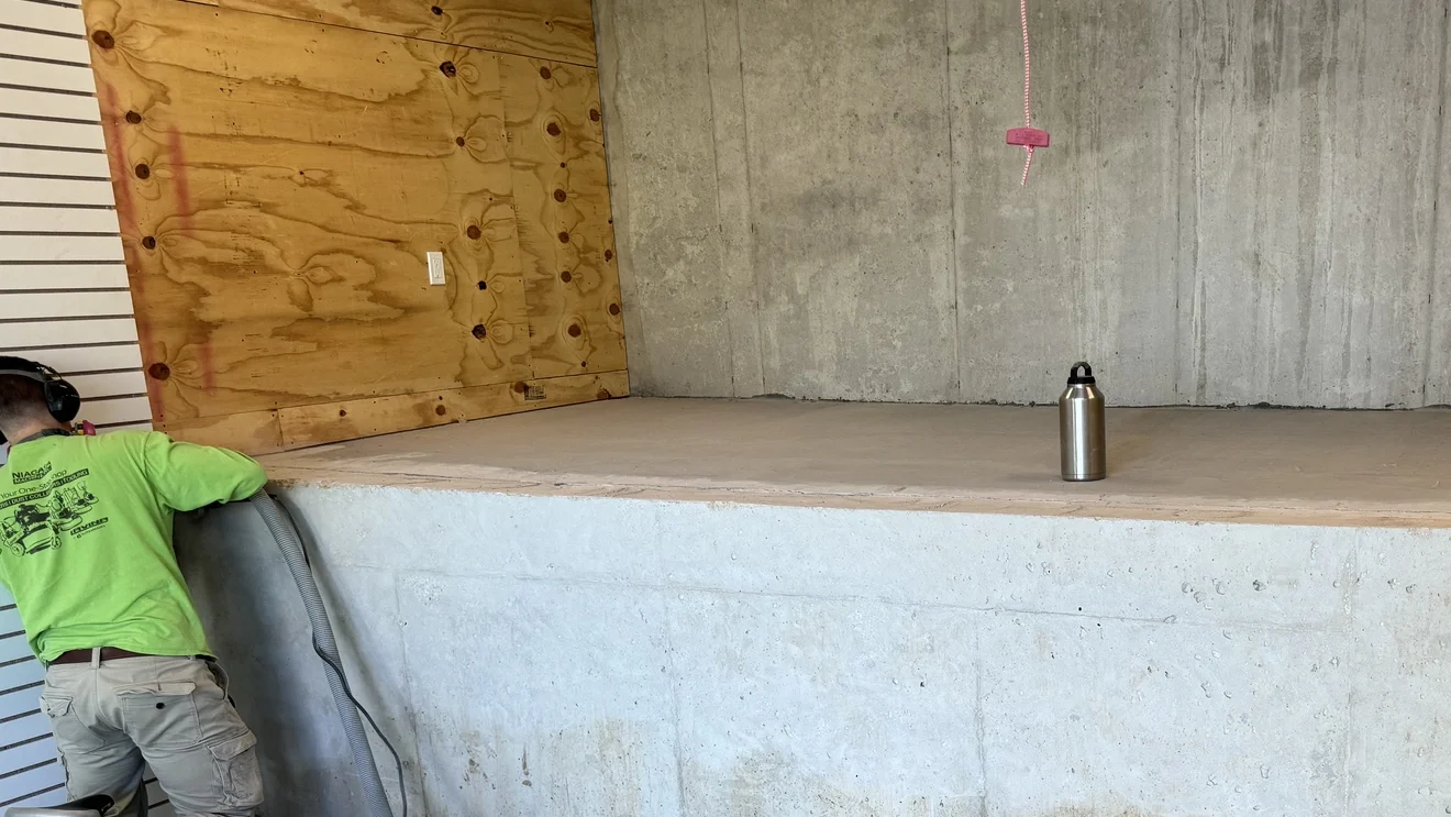 Worker preparing concrete floor for coating installation in commercial space with power tools and hoses