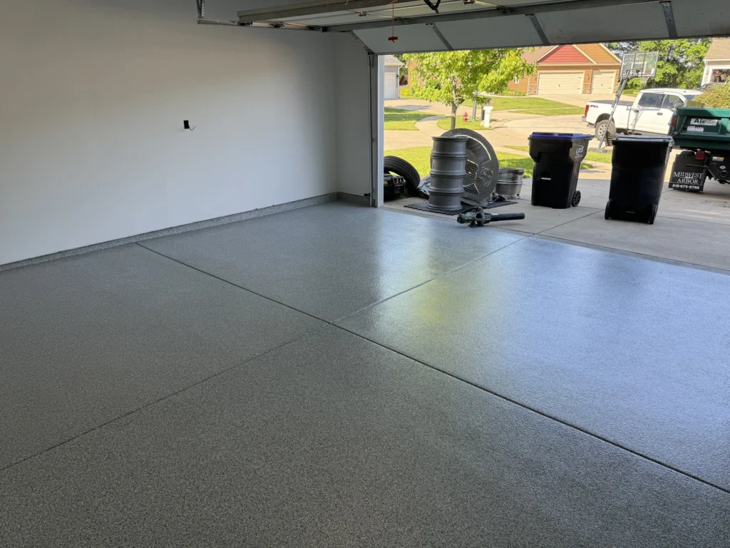 Gray epoxy coated garage floor with equipment and trash cans visible through the open garage door