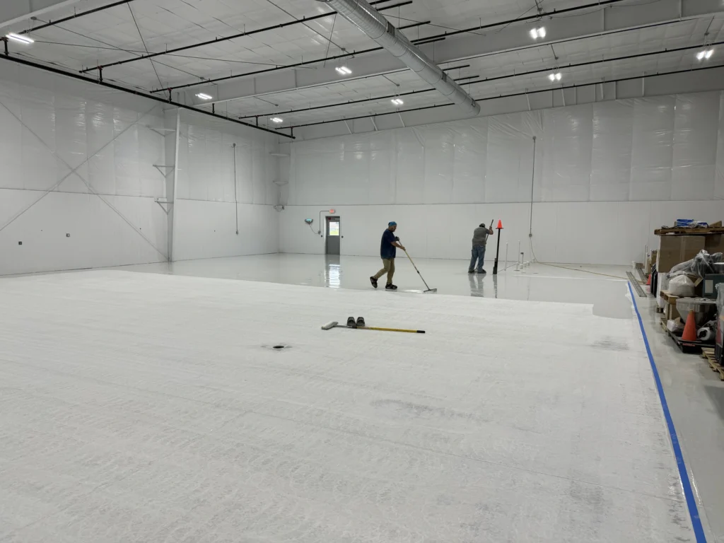 Workers installing white resinous floor coating in a large industrial warehouse with high ceilings and exposed ductwork