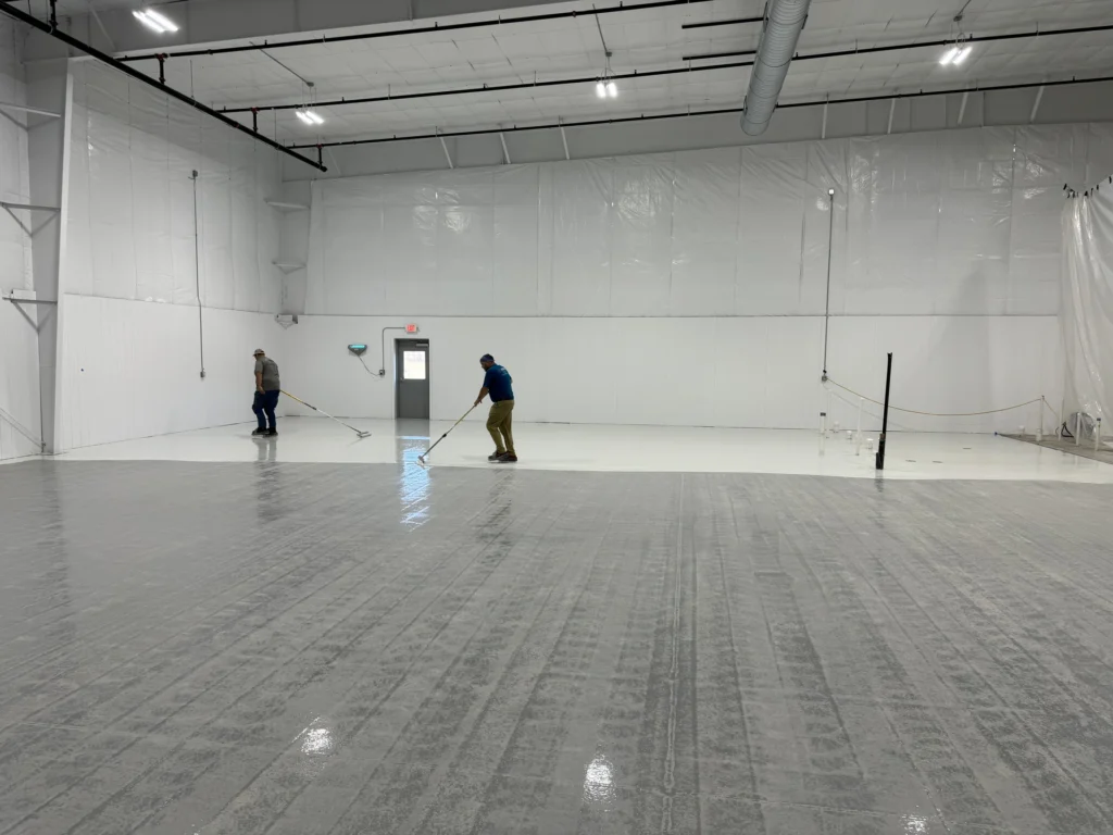 Workers installing a new floor coating in a large, empty warehouse with white walls and plastic sheeting